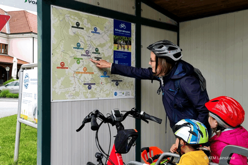 Frau und zwei Kinder mit Fahrradhelmen betrachten einen Radfahrplan mit ihren Rädern.