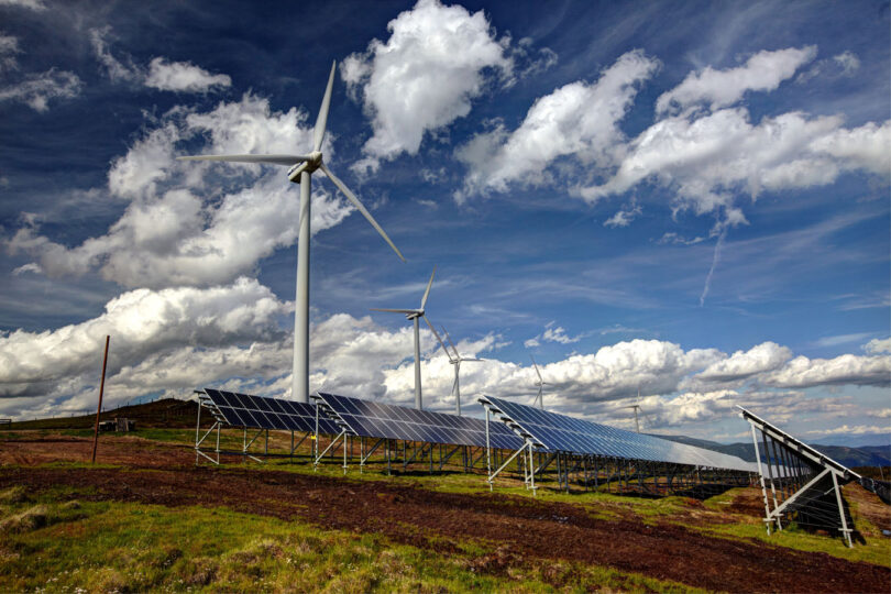 Mehrere Windräder und Solarpaneele stehen auf einer hügeligen Landschaft unter einem teils bewölkten Himmel. Die Kombination von Wind- und Solarenergieanlagen symbolisiert nachhaltige Stromerzeugung aus erneuerbaren Quellen.