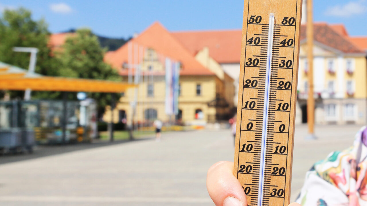 Eine Person hält ein Thermometer im Freien, das eine Temperatur von etwa 35 °C anzeigt. Im Hintergrund sind Gebäude und ein Platz bei sonnigem Wetter zu sehen.