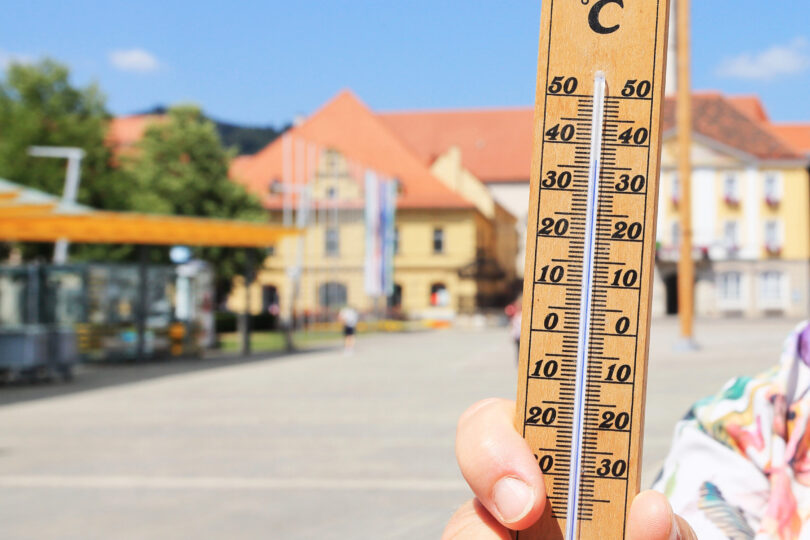 Eine Person hält ein Thermometer im Freien, das eine Temperatur von etwa 35 °C anzeigt. Im Hintergrund sind Gebäude und ein Platz bei sonnigem Wetter zu sehen.
