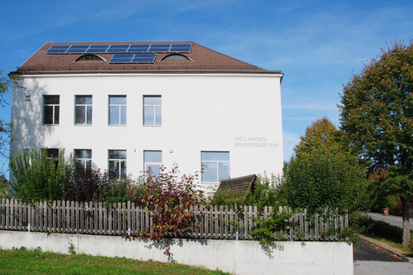 Außenansicht eines weißen Gebäudes mit der Aufschrift „NÖ LANDES-KINDERGARTEN“, großen Fenstern, Solarpaneelen auf dem Dach, einem Holzzaun und dicht bewachsener Gartenfläche im Vordergrund unter blauem Himmel.