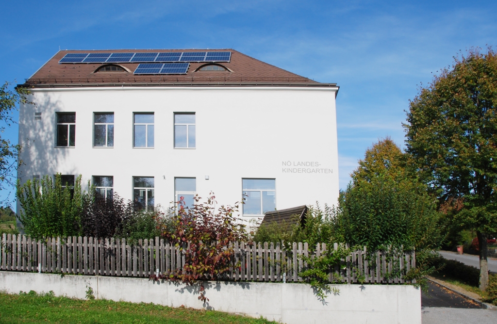 Außenansicht eines weißen Gebäudes mit der Aufschrift „NÖ LANDES-KINDERGARTEN“, großen Fenstern, Solarpaneelen auf dem Dach, einem Holzzaun und dicht bewachsener Gartenfläche im Vordergrund unter blauem Himmel.