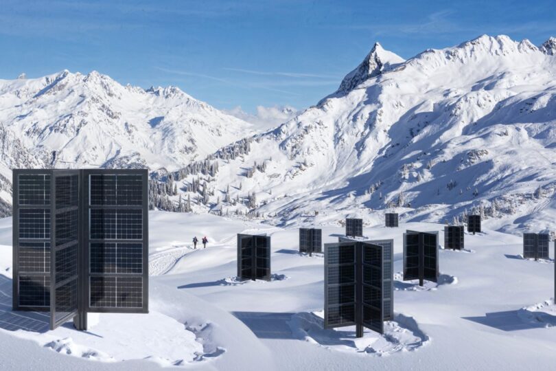 Vertikale Solarpanels in einer schneebedeckten Berglandschaft, umgeben von majestätischen Gipfeln unter einem klaren blauen Himmel. Im Hintergrund sind zwei Menschen auf einem verschneiten Pfad unterwegs, was die Verbindung von erneuerbarer Energie und Natur in extremen Klimazonen betont.