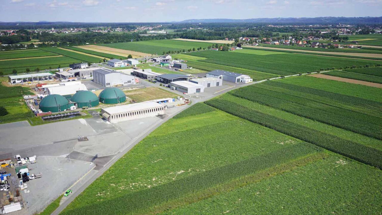 Luftaufnahme einer modernen Biogasanlage inmitten landwirtschaftlicher Felder. Mehrere große grüne Kuppeln sind sichtbar, umgeben von Industriegebäuden und befestigten Flächen. Im Hintergrund erstrecken sich Felder, kleine Ortschaften und eine hügelige Landschaft unter einem leicht bewölkten Himmel.