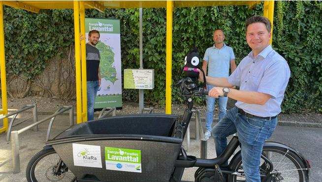 Ein Mann mit einem Fahrradkarren steht unter einem gelben Unterstand, neben einer Karte und Informationsplakaten über Lavanttaler Energieprojekte.