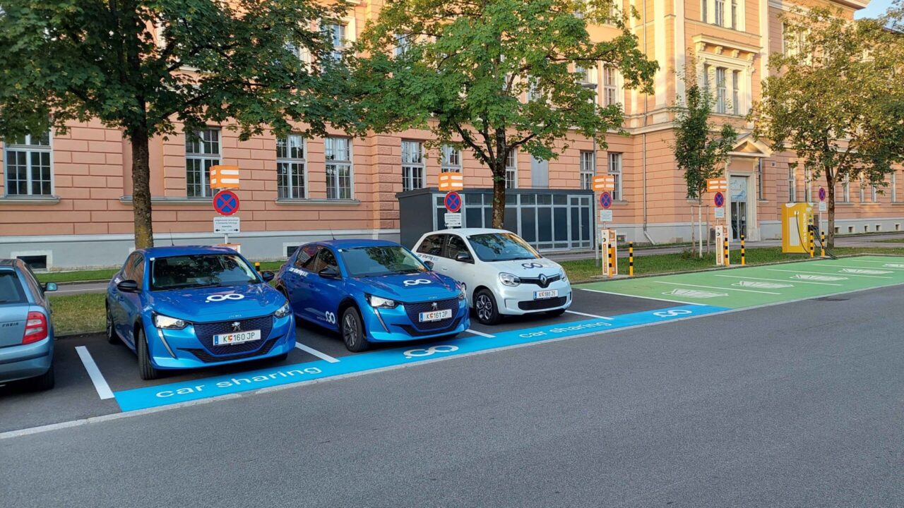 Drei Parkende E-Autos an Ladestationen. Im Hintergrund Bäume, ein Energiespeicher und ein Gebäude.