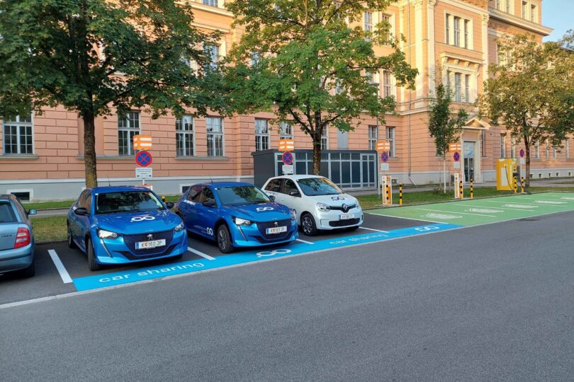 Drei Parkende E-Autos an Ladestationen. Im Hintergrund Bäume, ein Energiespeicher und ein Gebäude.