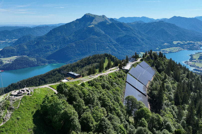 Luftaufnahme von Bergen mit einem See, Straßen, Gebäuden und Solaranlagen inmitten von Grünflächen.