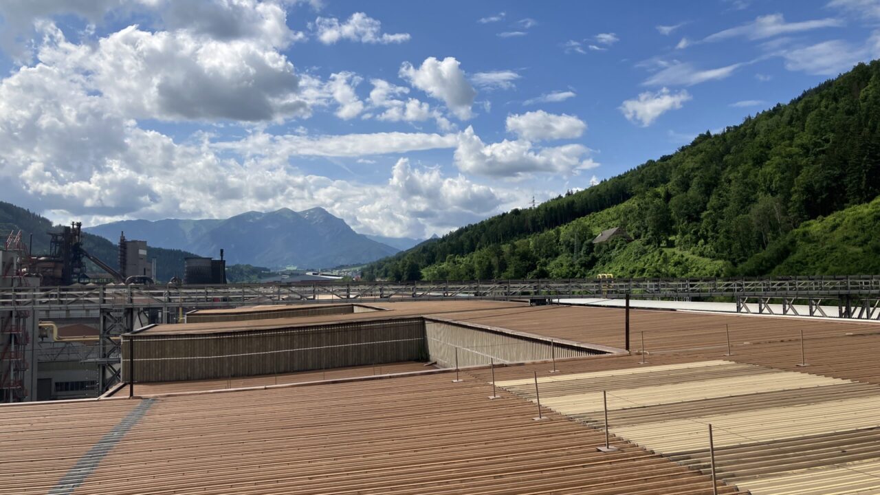 Industriegelände mit großen Holzschornsteinen, umgeben von grünen Bergen unter einem bewölkten Himmel.