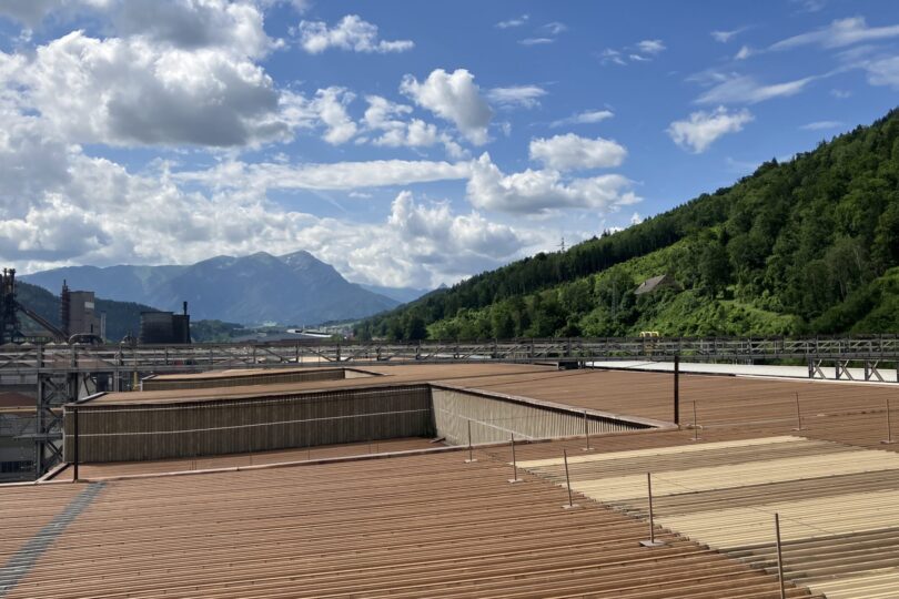 Industriegelände mit großen Holzschornsteinen, umgeben von grünen Bergen unter einem bewölkten Himmel.