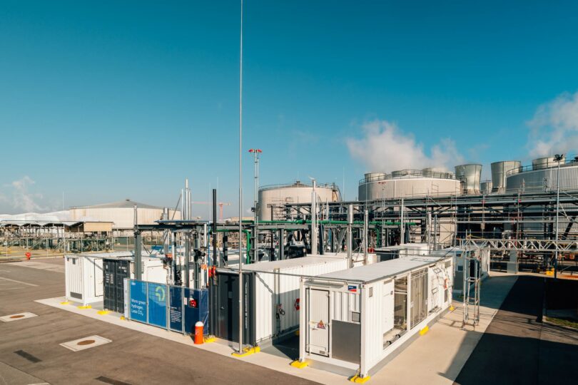Industrieanlage mit mehreren großen zylindrischen Tanks und weißen Containern auf Asphaltfläche unter blauem Himmel.