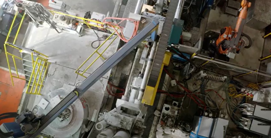 Industrieanlage mit komplexem Maschinenaufbau in einer Produktionshalle, aufgenommen aus der Vogelperspektive. Ein großer Roboterarm in Orange befindet sich in der oberen rechten Ecke, während Förderbänder, Metallplattformen mit gelben Geländern und zahlreiche Kabelstränge das Bild dominieren.