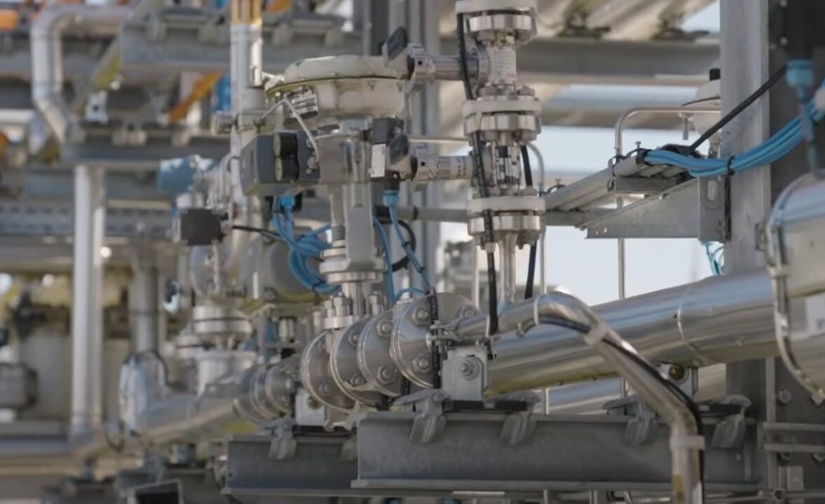 Ein komplexes industrielles Rohrleitungssystem mit verschiedenen Ventilen, Metallrohren und blauen Kabelverbindungen. Die glänzenden Rohre reflektieren das Sonnenlicht, während im Hintergrund weitere Rohrleitungen und industrielle Strukturen sichtbar sind.