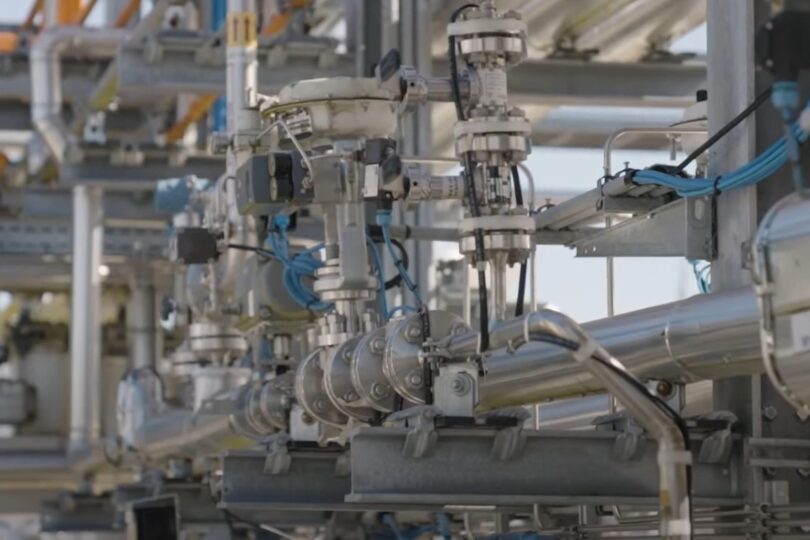 Ein komplexes industrielles Rohrleitungssystem mit verschiedenen Ventilen, Metallrohren und blauen Kabelverbindungen. Die glänzenden Rohre reflektieren das Sonnenlicht, während im Hintergrund weitere Rohrleitungen und industrielle Strukturen sichtbar sind.