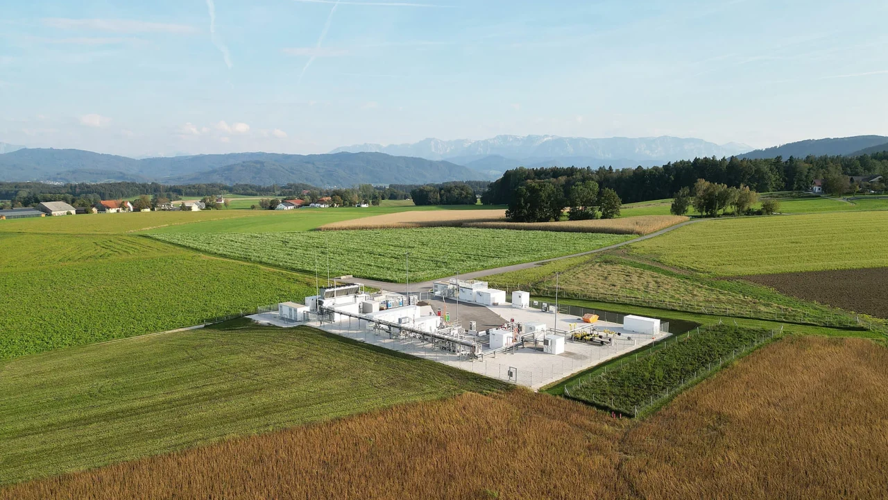 Luftaufnahme einer Industrieanlage mit mehreren weißen Tanks und Gebäuden, umgeben von landwirtschaftlichen Feldern und Bergen im Hintergrund.