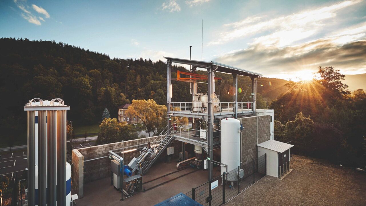 Industrielle Anlage mit mehreren silbernen Tanks und Rohren vor bewaldetem Hügel und Sonnenuntergang.