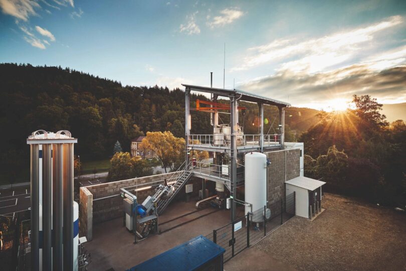 Industrielle Anlage mit mehreren silbernen Tanks und Rohren vor bewaldetem Hügel und Sonnenuntergang.